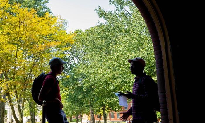 The outline of two students standing in a corridor in Seaver Hall with a bright outdoor background of green trees on the Harvard Quad. 