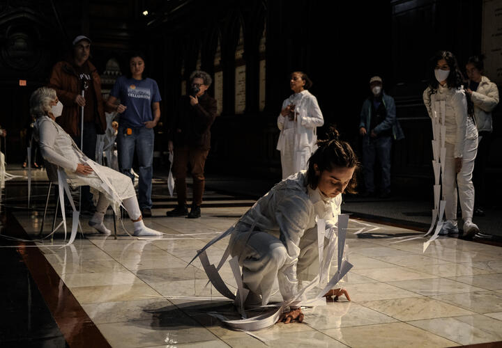 Four dancers wearing white whimsical paper sculpture across their bodies, one reclining in a chair, one kneeling on ground, two standing straight