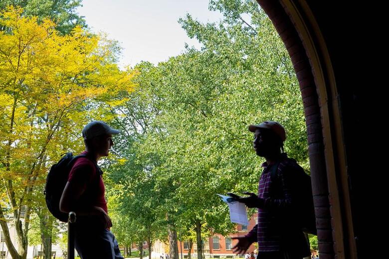 The outline of two students standing in a corridor in Seaver Hall with a bright outdoor background of green trees on the Harvard Quad. 