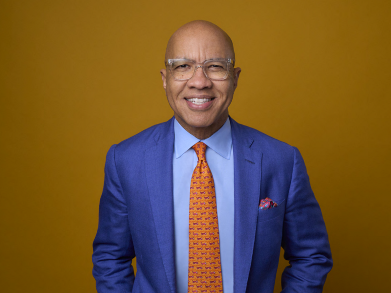 A waist up photo of Darren Walker, smiling in a bright blue suit and orange tie.
