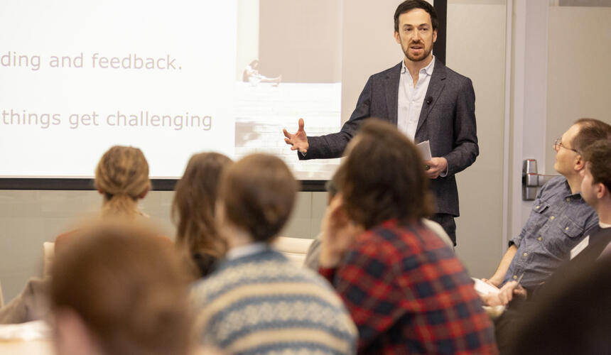 A teacher stands in front of a classroom with a lesson projected on the wall behind. 