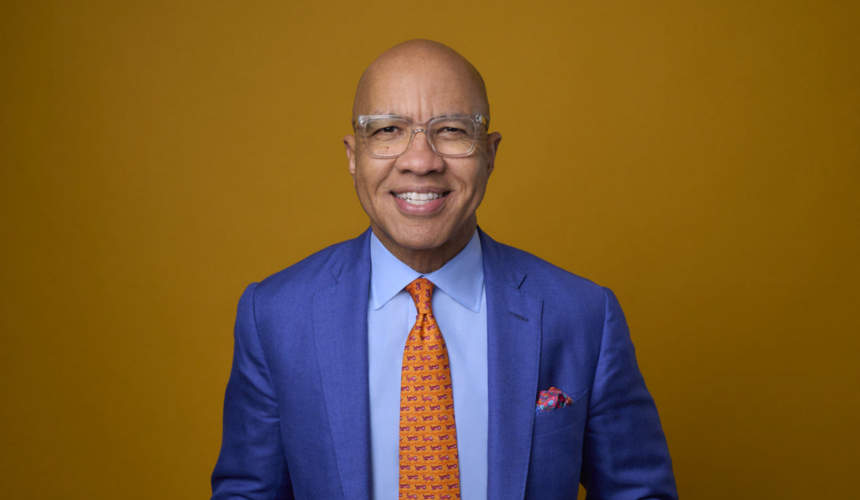 A waist up photo of Darren Walker, smiling in a bright blue suit and orange tie.