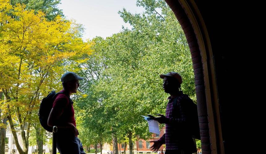 The outline of two students standing in a corridor in Seaver Hall with a bright outdoor background of green trees on the Harvard Quad. 